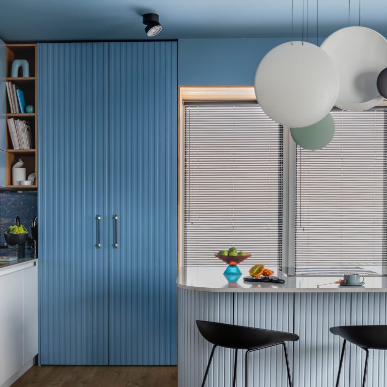 Creative composition of kitchen interior with marble kitchen island, blue kitchen furnitures, black chokers, modern lamp, bowl with fruits, big window and personal accessories.